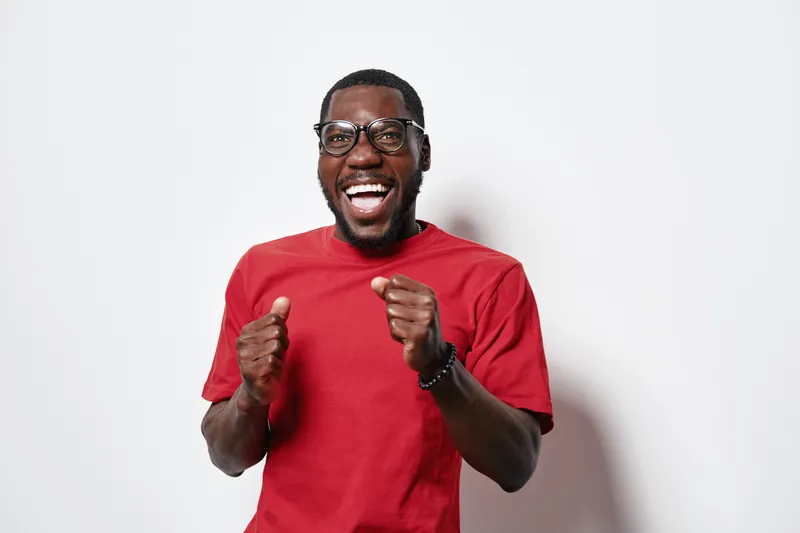 African American man with dark skin wearing glasses and red t shirt smiling and celebrating with fists up on white background. Happy male portrait with positive emotion.
