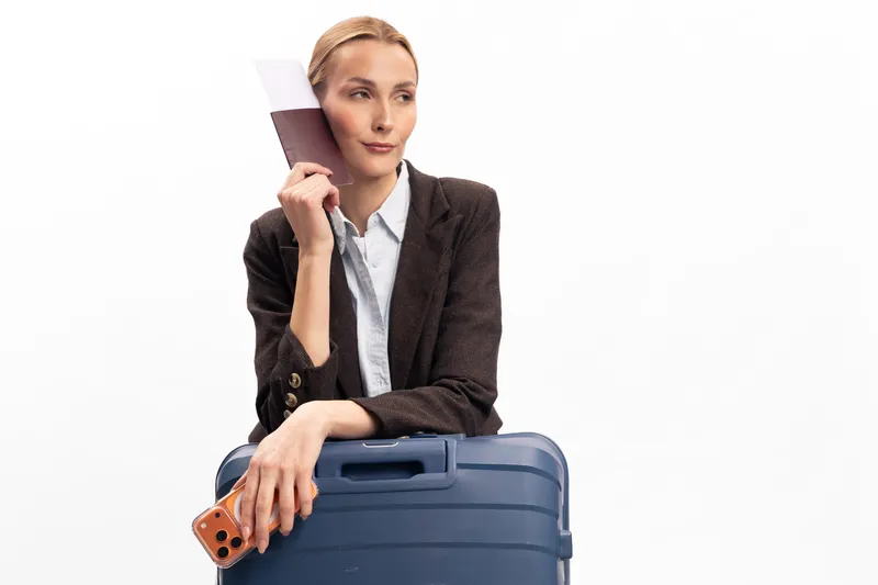 A thoughtful woman holds a color swatch near her face while leaning on a large blue suitcase, dressed in a blazer and white shirt, isolated on white background. Travel and decision concept.