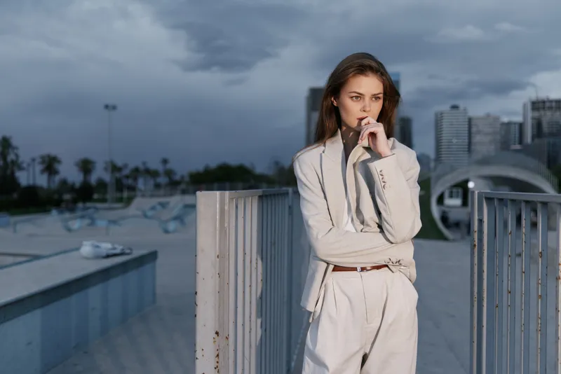 Thoughtful woman in beige business suit standing outdoors with cityscape and cloudy sky, expressing focus and contemplation