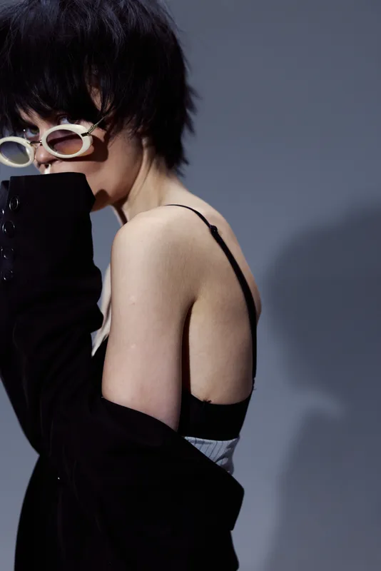 Stylish woman with short black hair wearing sunglasses and a black jacket, posing with mysterious emotion against gray background.