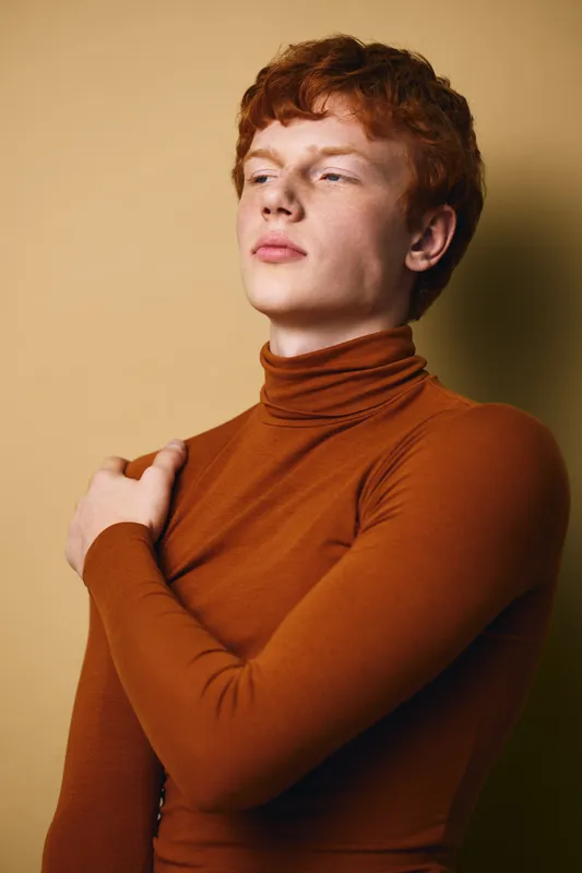 Young man with red hair wearing brown turtleneck posed with hand on shoulder against beige background. Fashion portrait, casual style, confident expression and soft lighting focus.
