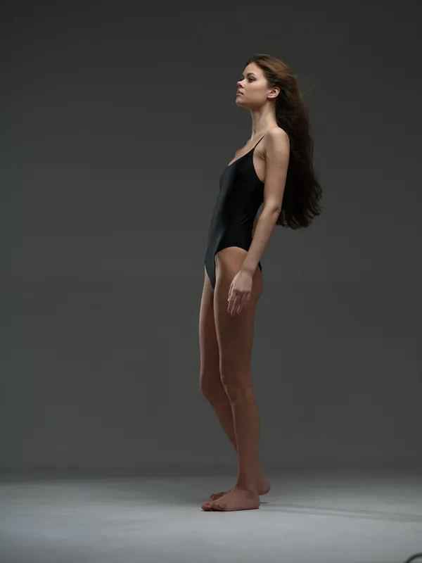 Tall woman with long hair standing barefoot in black bodysuit against plain gray background, calm expression, studio lighting.