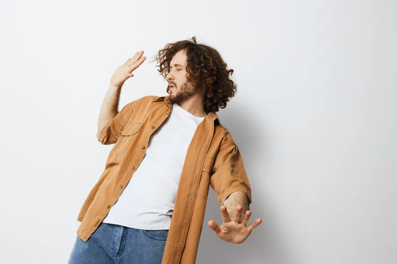 Young man with curly hair expressing joy in a casual outfit, wearing a beige shirt and white t shirt, against a light background, capturing a feeling of happiness and spontaneity