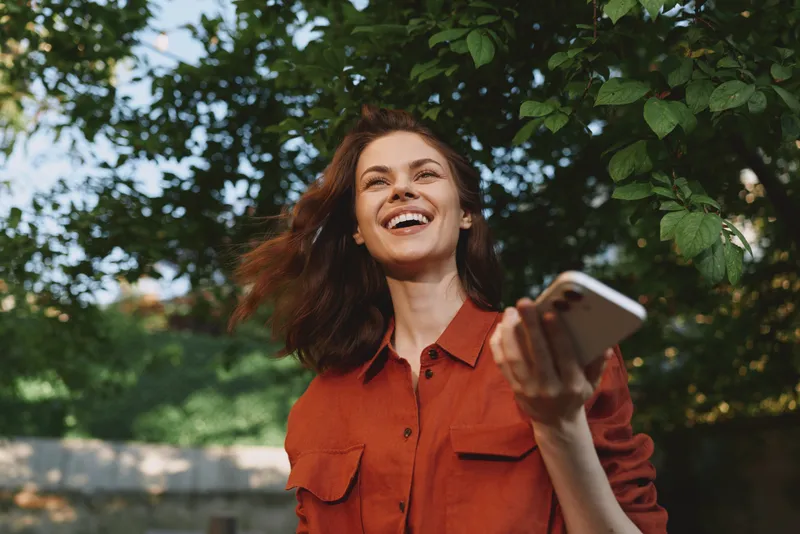 Smiling woman outdoors wearing a red shirt, holding a smartphone. Close-up on leafy background. Concept of lifestyle, connection, and modern daily moments. Soft daylight creates a warm, joyful mood