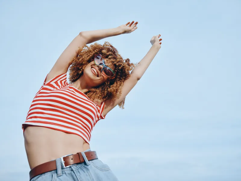 Joyful woman with curly hair dancing under a clear blue sky, wearing a red and white striped crop top and denim jeans, capturing carefree summer energy and movement.