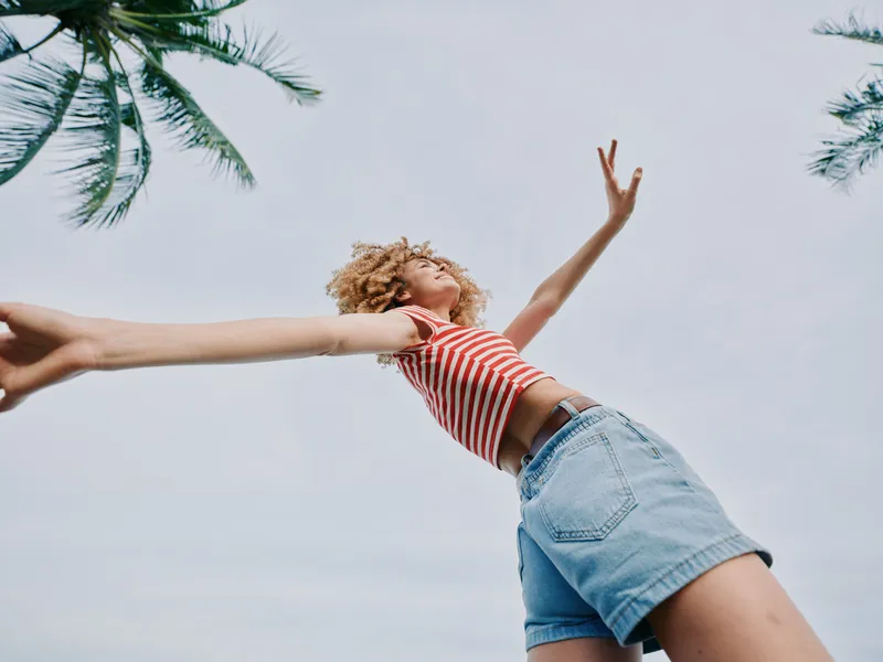A dynamic figure leaps against a clear sky, arms open wide and a striped top paired with denim shorts, capturing carefree summer energy and vibrant motion.