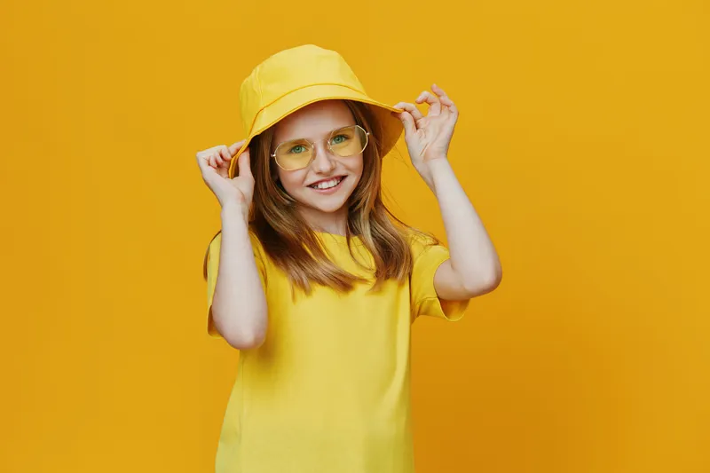 Cute girl wearing a yellow outfit with a matching hat and glasses against a vibrant yellow background, radiating joy and summer vibes