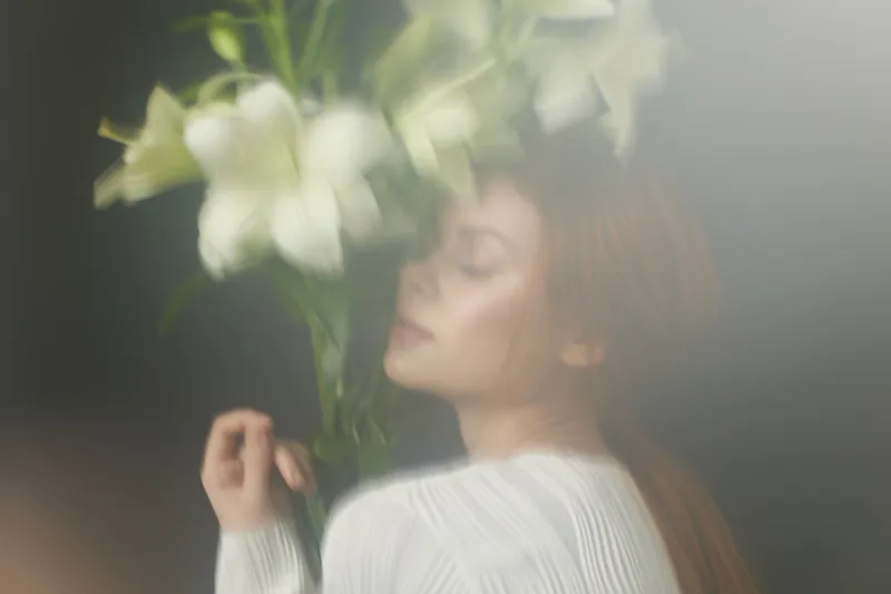 Young woman with long hair embraces white lilies, conveying serenity and grace against a soft, blurred background, perfect for wellness and floral themes
