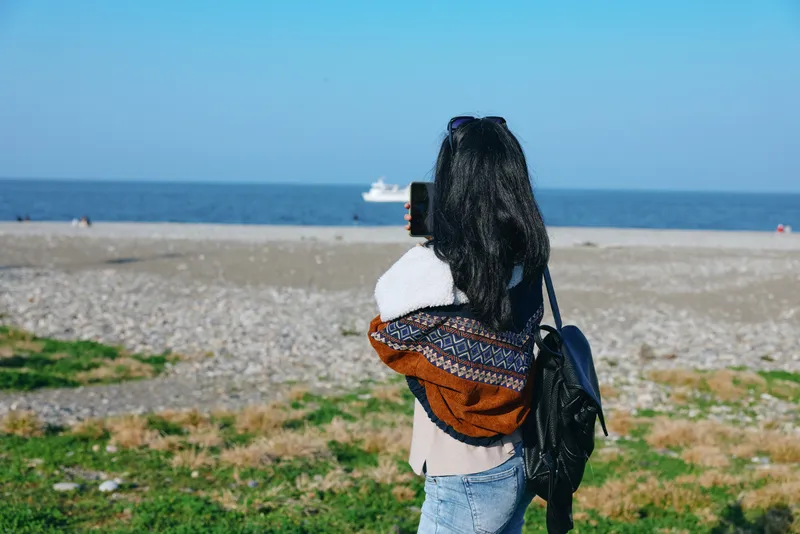 Woman on a sandy beach with backpack gazing at the sea, casual jacket and scarf, exploring coastal landscape and outdoor travel moment