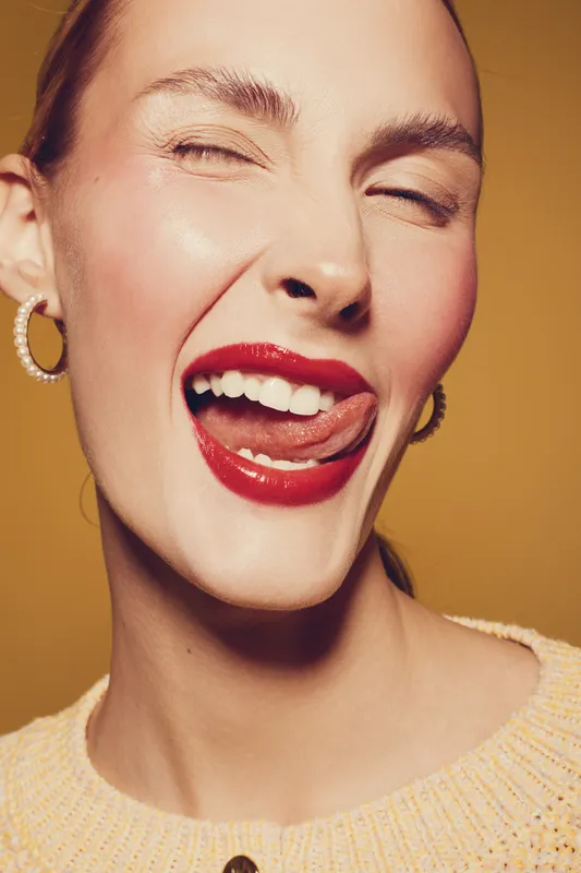 Close-up of a young woman with red lipstick winking and sticking out her tongue. She wears pearl earrings and a knitted sweater on a warm beige background. Playful and confident mood.
