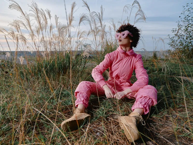 Outdoor person wearing pink fashion sits among tall grasses in a field, relaxed pose with boots, wide sky, natural setting, calm mood, candid lifestyle scene for travel or editorial use