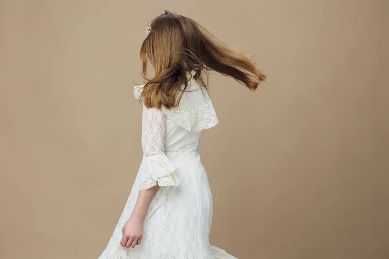 Young girl in a flowing white dress, spinning gracefully against a soft beige background, showcasing joy and freedom in a whimsical moment of childhood