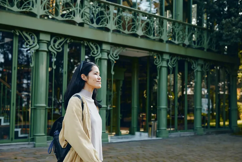 Woman with backpack strolling outdoors beside ornate green arches, wearing a light jacket, calm smile, capturing travel mood, urban leisure and outdoor style in a sunlit park setting