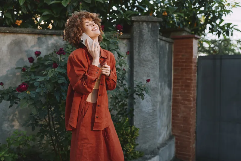 One young woman stands outdoors in an orange cardigan, smiling softly as sunlight highlights her curly hair and relaxed pose in a blooming garden setting