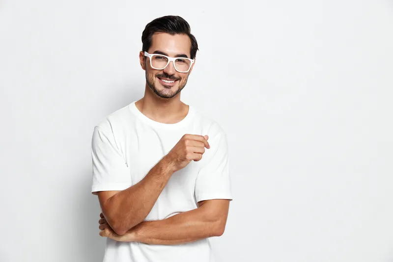 Smiling man with glasses dressed in a casual white t-shirt stands confidently, posing with a charming smile against a light solid background. Lifestyle concept.