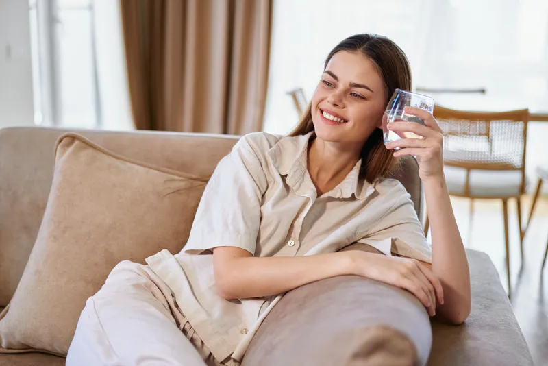 Woman on sofa at home with a smile, relaxed on a couch in a bright livingroom, drinking water from a glass and enjoying quiet relaxation, feeling calm and happy in casual clothes.