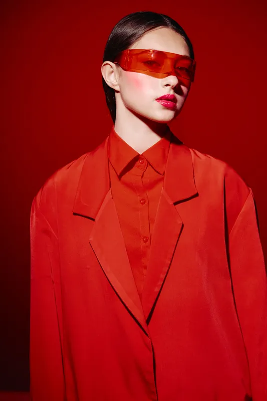 Young woman wearing oversized red blazer and futuristic tinted glasses with sleek hair in matching red background. Concept of avant-garde beauty fashion and creative style.