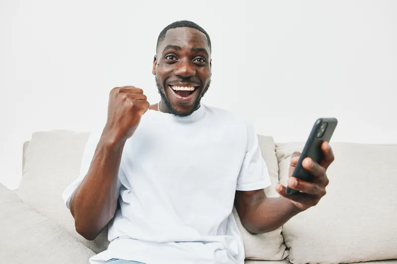 Excited young African man celebrating victory while using smartphone on a light background in a casual outfit
