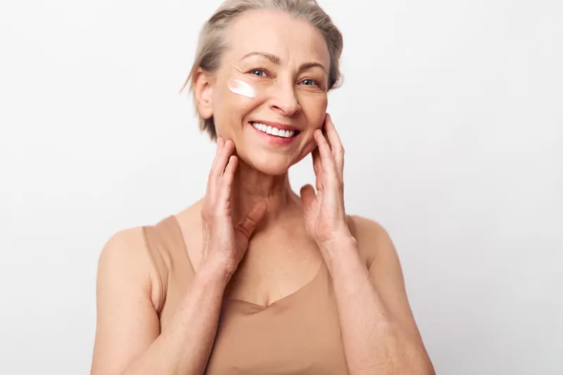 Smiling mature woman with short hair applies white facial cream on cheeks, showing natural skin care and beauty routine. Bright studio shot with soft lighting and beige top. Concept of healthy aging.