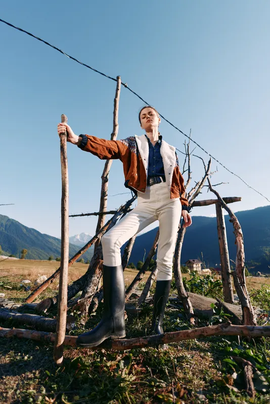 Woman in rubber boots holding a wooden stick by a rustic fence, wearing brown jacket and white trousers in countryside, standing confident with mountain valley and clear blue sky.