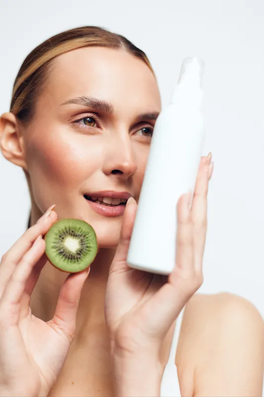 Young woman holding kiwi slice and white cosmetic bottle, clear skin, natural beauty, skincare concept, bright studio background, fresh healthy look, minimal makeup