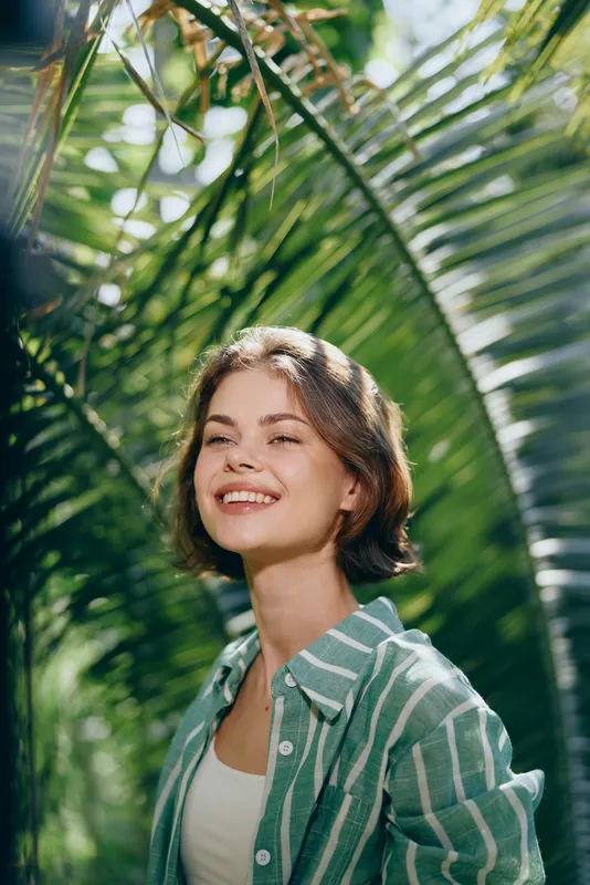 Portrait of a cheerful woman in a striped green shirt beneath tropical palm leaves, basking in sunlight.