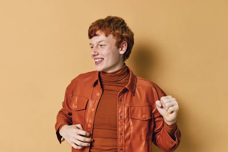 Young man with red hair wearing orange jacket and turtleneck dancing and smiling looking to side on plain beige background in studio. Casual style, happiness and movement portrait.