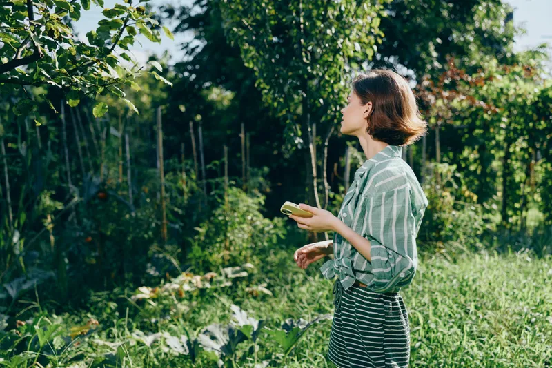 A woman standing in a lush garden lights up with sun as she holds a smartphone, surrounded by green leaves and fresh flowers.