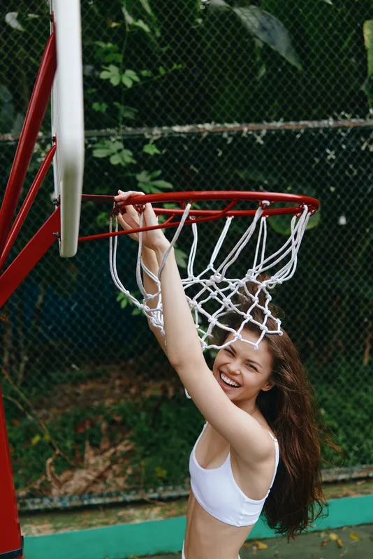 Joyful young woman in sporty attire smiling while hanging from a basketball hoop outdoors, capturing the essence of fun and athleticism in a vibrant setting