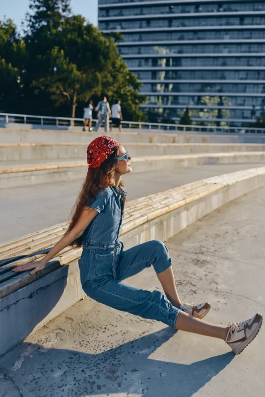 Young woman sitting outdoors on a sunny day wearing denim jumpsuit, red bandana, sunglasses, and sandals enjoying urban background and warm weather.