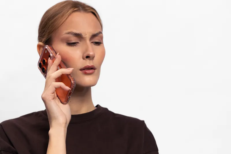 Young woman in black shirt looks confused while holding smartphone to ear, isolated on white background. Concept of communication, technology, and uncertain conversation.