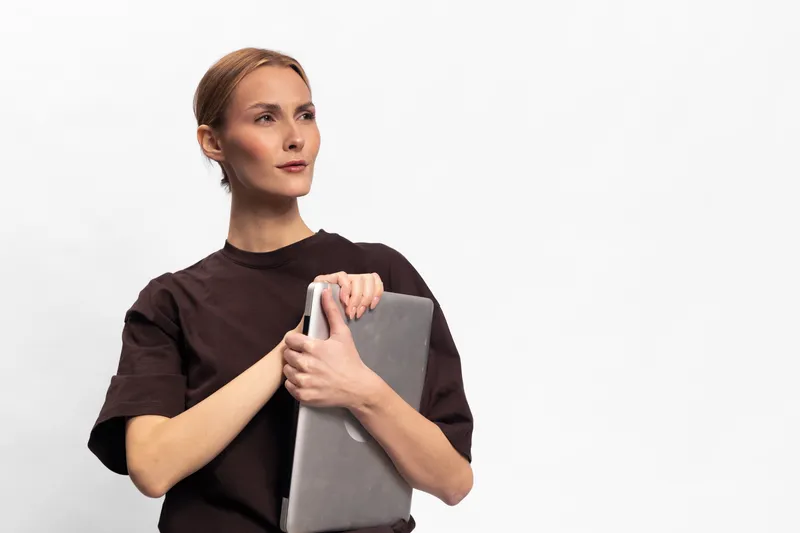 Confident woman in dark casual shirt holding a laptop, looking away thoughtfully on white background. Concept for technology, business, and modern lifestyle.