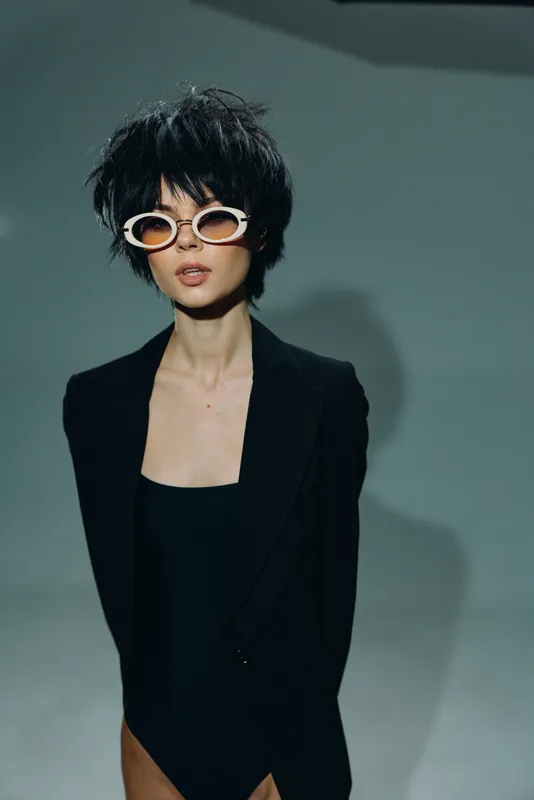 Stylish woman with short hair wearing white sunglasses and black blazer posing confidently in studio lighting.
