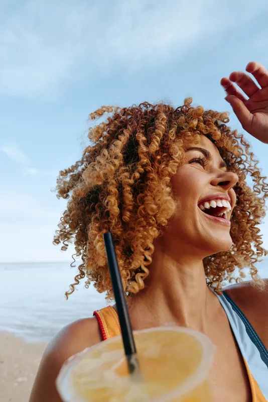 Woman with curly hair on beach, laughing in sunlight, holding a hand up, sipping a summer drink with lemon, ocean horizon and waves behind, joyful relaxed mood