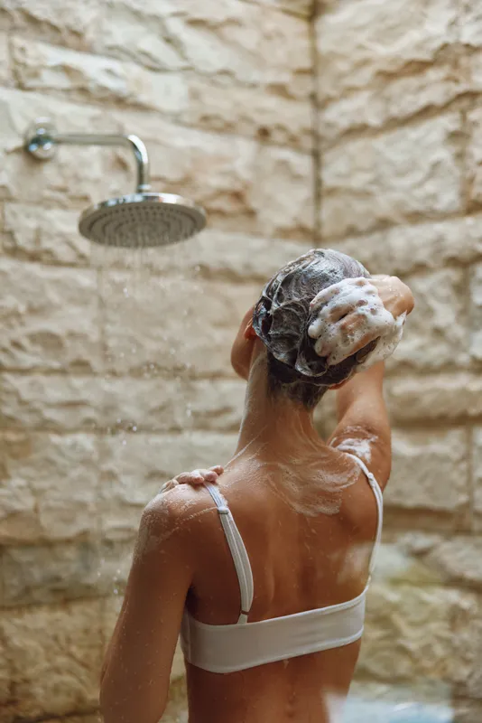 woman washing hair under rain shower with foam, soft skin care, morning routine in warm stone bathroom, refreshing and calming atmosphere