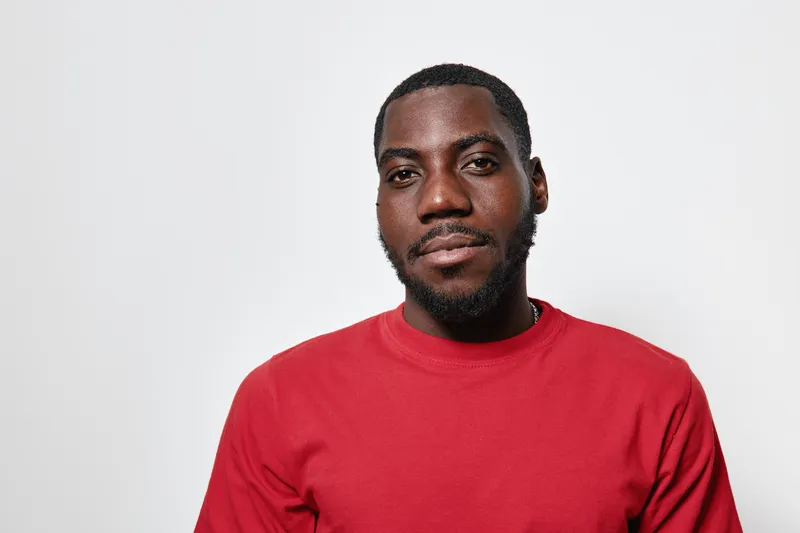 African American man with dark skin wearing red sweater looking at camera with calm expression on white background. Portrait of confident black male with short hair and beard.