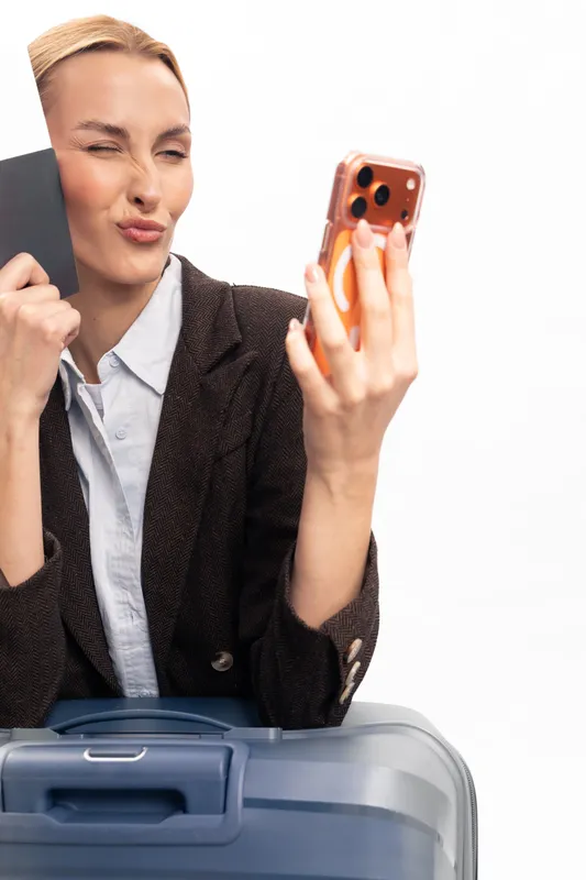 Young woman in a brown jacket makes a selfie with a phone while holding a passport near a blue suitcase. Travel concept with excitement and preparation before trip.