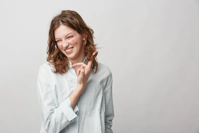 Young woman with curly hair wearing white shirt smiles and holds a small object between fingers on light background. Concept of happiness, casual style, and positive emotions.