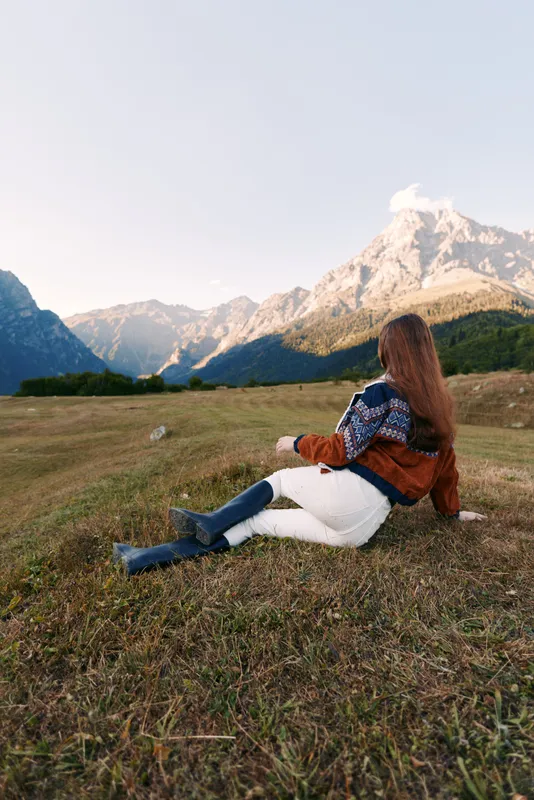 Woman in sweater and boots sits on a mountain meadow, looking at alpine mountains and vast nature landscape during autumn travel, relaxing solitude and scenic outdoor view.
