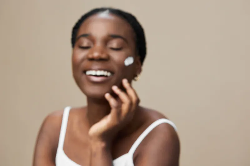 Joyful African woman applying skincare product with a natural smile in a soft, neutral toned background, showcasing self care and beauty concepts