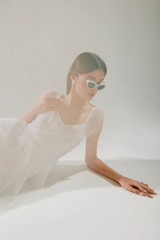 Young woman in a white sheer dress wearing white sunglasses lies on a light surface. Soft lighting highlights her beauty and fashion style in a minimalistic setting.