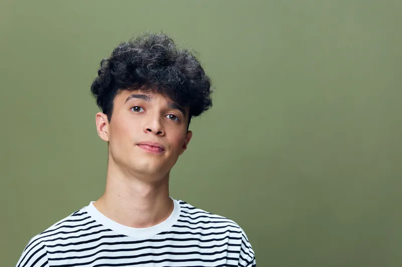young man with curly hair wearing striped t-shirt poses against plain olive background, casual style with soft natural lighting in studio setting