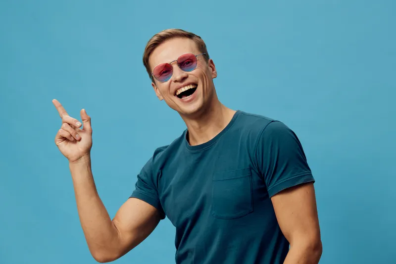 smiling young caucasian man wear sunglasses teal t-shirt point finger look camera isolated blue background casual style digital nomad concept