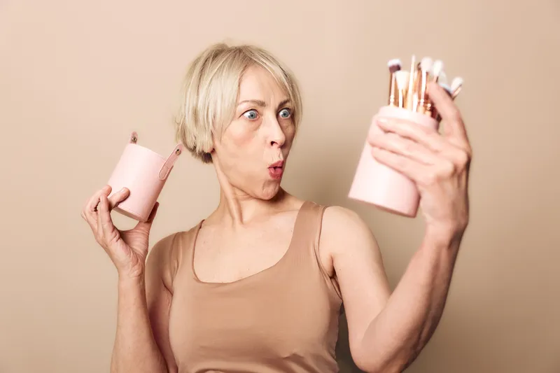 Surprised middle aged woman with short blonde hair holds makeup brushes and container, looking at them with wide eyes and open mouth. Concept of beauty routine, cosmetics, and skincare.