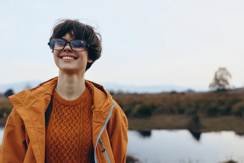 Woman lifestyle outdoors, smiling in glasses, wearing a brown jacket and orange sweater, standing by a calm lake at dusk, soft wind, natural countryside background, gentle sunset light calm
