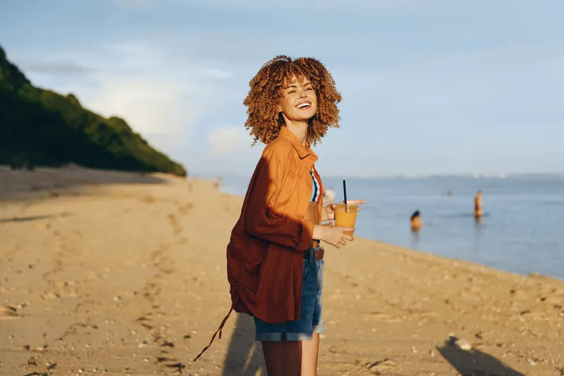 Young woman with curly hair enjoys a refreshing drink while standing on a sunny beach with a relaxed smile and casual summer outfit.