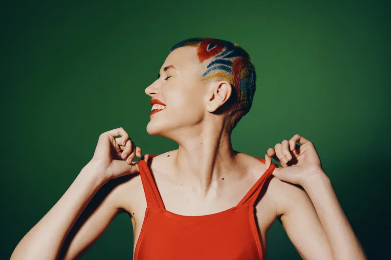Smiling woman with artistic shaved hairstyle and bright red lipstick wearing red sleeveless top on green background, expressing joy and confidence with eyes closed.