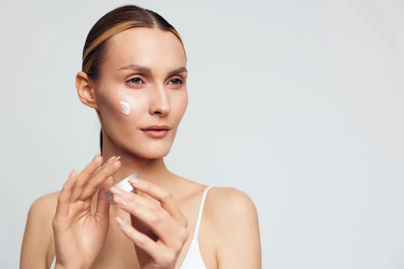 Woman with tied hair applying white face cream on cheek, holding jar, looking away, on light background. Concept of skincare, beauty routine, and natural wellness.