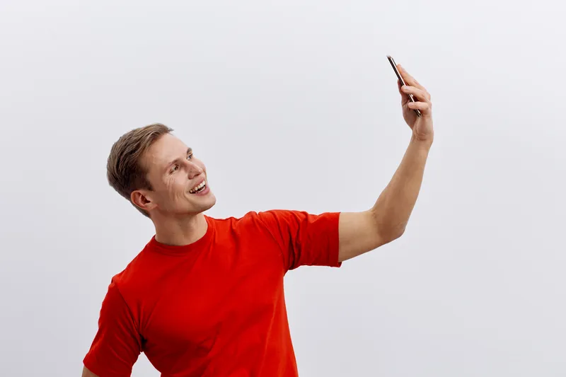 Smiling young man taking a selfie with smartphone, casual clothing, red t-shirt, cheerful expression, white background, outdoor activity, lifestyle concept