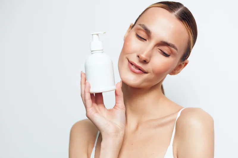 A smiling young woman with clear skin holds a white cosmetic bottle close to her face against a plain white background. Concept for skincare, beauty, and personal care.